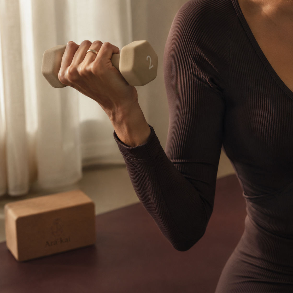 A woman in a ribbed long-sleeve dark plum activewear top lifts a beige hex dumbbell on a yoga mat, with a wooden yoga block in the background.